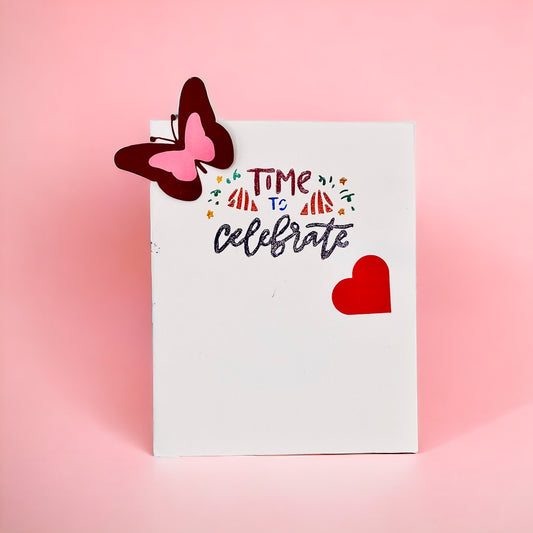 A card titled “ Time to Celebrate” with a pop-out butterfly and a red heart sticker on a pink background 