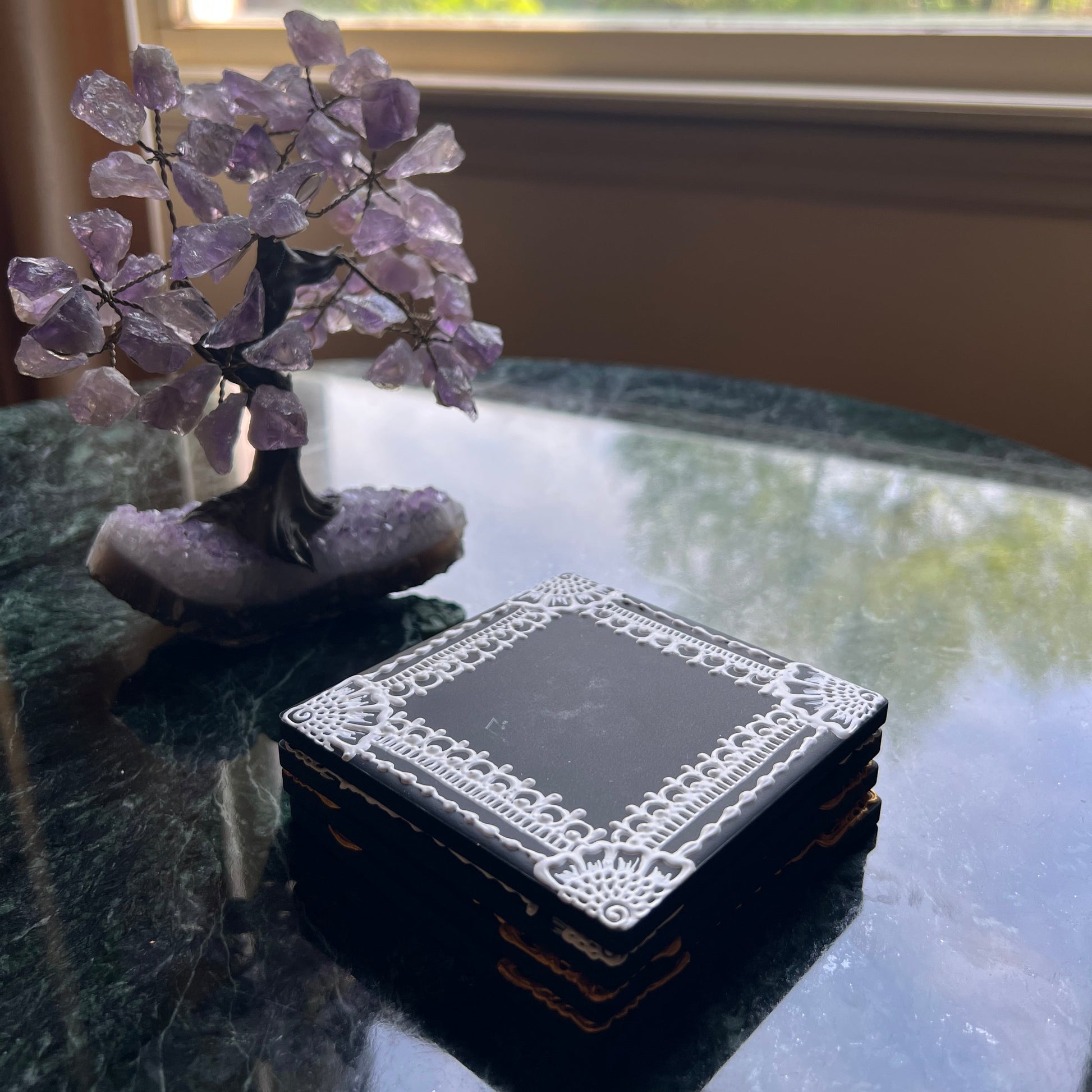 Stack of hand-painted ceramic coasters with white henna border on black tile, styled on glass table with amethyst tree decor